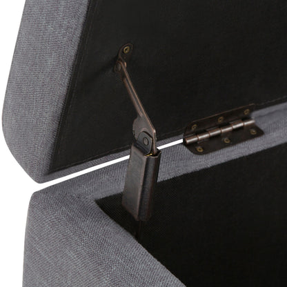 Close-up of the Hamilton - Upholstered Storage Ottoman in grey with its lid open, revealing the metallic hinge and support mechanism inside. The fabric texture and hardware details are clearly visible.