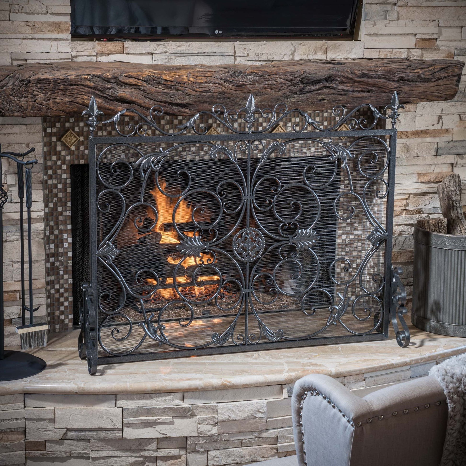 A lit fireplace features the Darcie Wrought Fireplace Screen with an ornate design, set in a stone mantel with a thick wooden beam and TV above. A chair and metal bin sit nearby.
