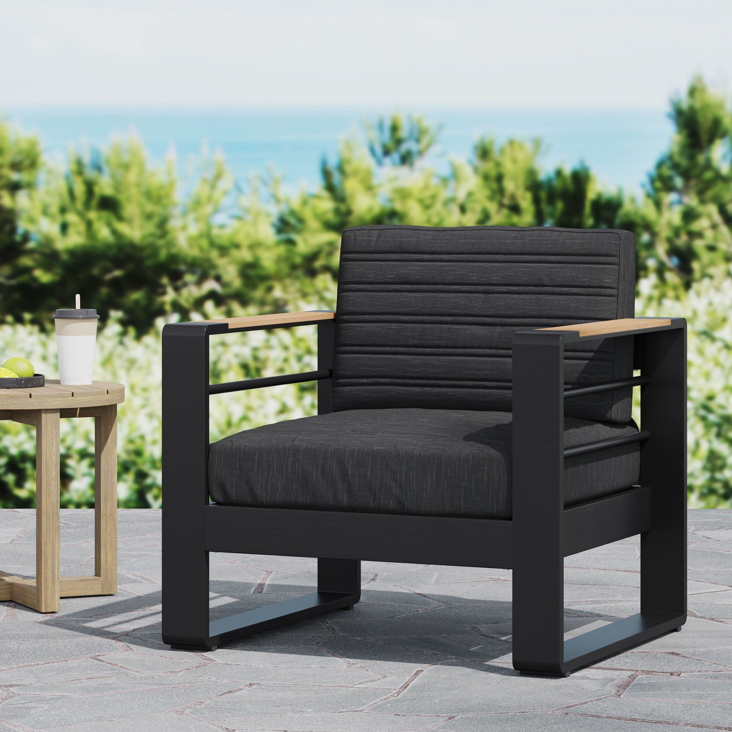 The Giovanna - Club Chair Set with Aluminum Frame features a modern black outdoor armchair with charcoal cushions on a stone patio, beside a small wooden table and coffee cup, with greenery and the blurred ocean in the background.