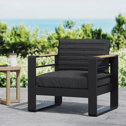 The Giovanna - Club Chair Set with Aluminum Frame features a modern black outdoor armchair with charcoal cushions on a stone patio, beside a small wooden table and coffee cup, with greenery and the blurred ocean in the background.