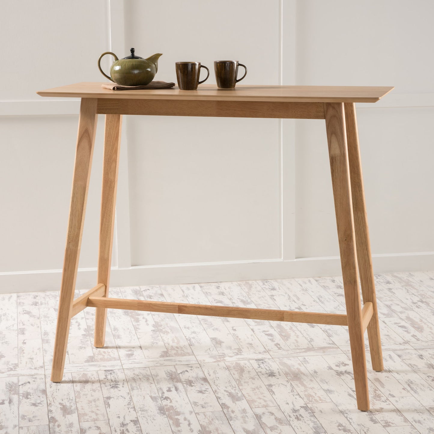 A Bar Height Dining Table With Splayed Legs And Elongated Rectangular Top displays a green teapot and two matching cups, set against a white wall and a light-colored wooden floor.