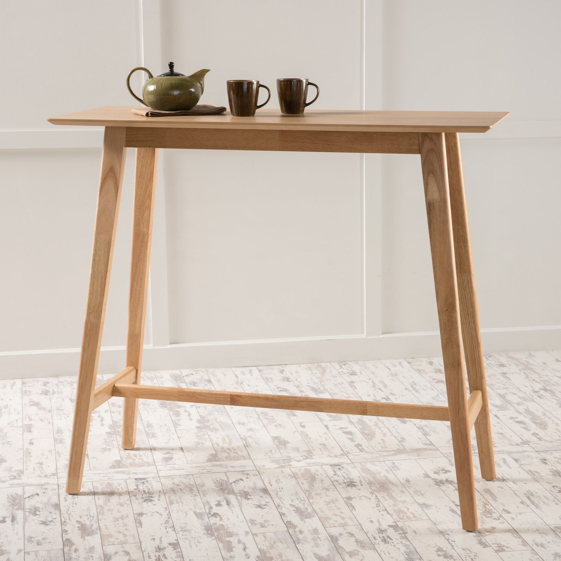 A Bar Height Dining Table With Splayed Legs And Elongated Rectangular Top displays a green teapot and two matching cups, set against a white wall and a light-colored wooden floor.