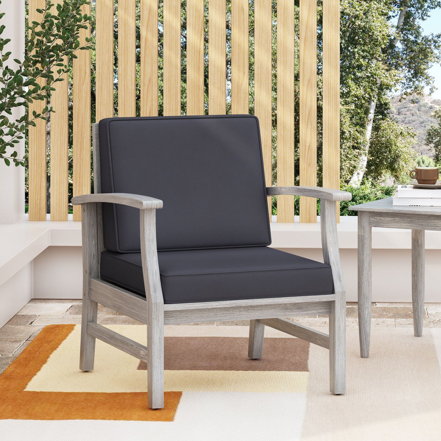 The Perla Contemporary Club Chair with water-resistant cushions, featuring a gray frame and dark gray cushions, sits on a patio rug beside a small table, with wooden slats and greenery in the background.
