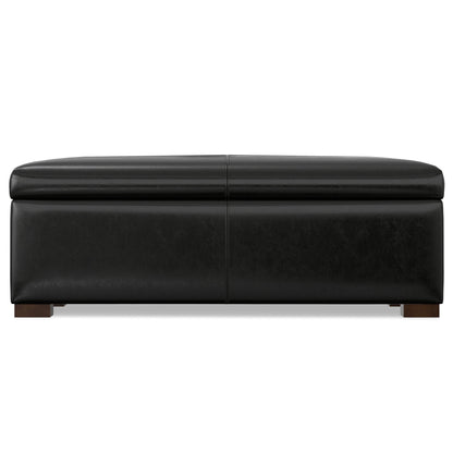 The Gabbie Coffee Table Upholstered Storage Ottoman features a rectangular black leather design with a cushioned top and short wooden legs, shown from the front.