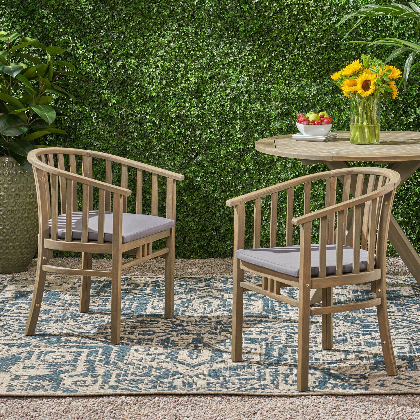 Two Outdoor Wooden Dining Chairs With Cushions (Set of 2) with gray cushions are arranged on a patterned outdoor rug near a round wooden table topped with fruit and sunflowers, surrounded by lush greenery and a leafy hedge backdrop.