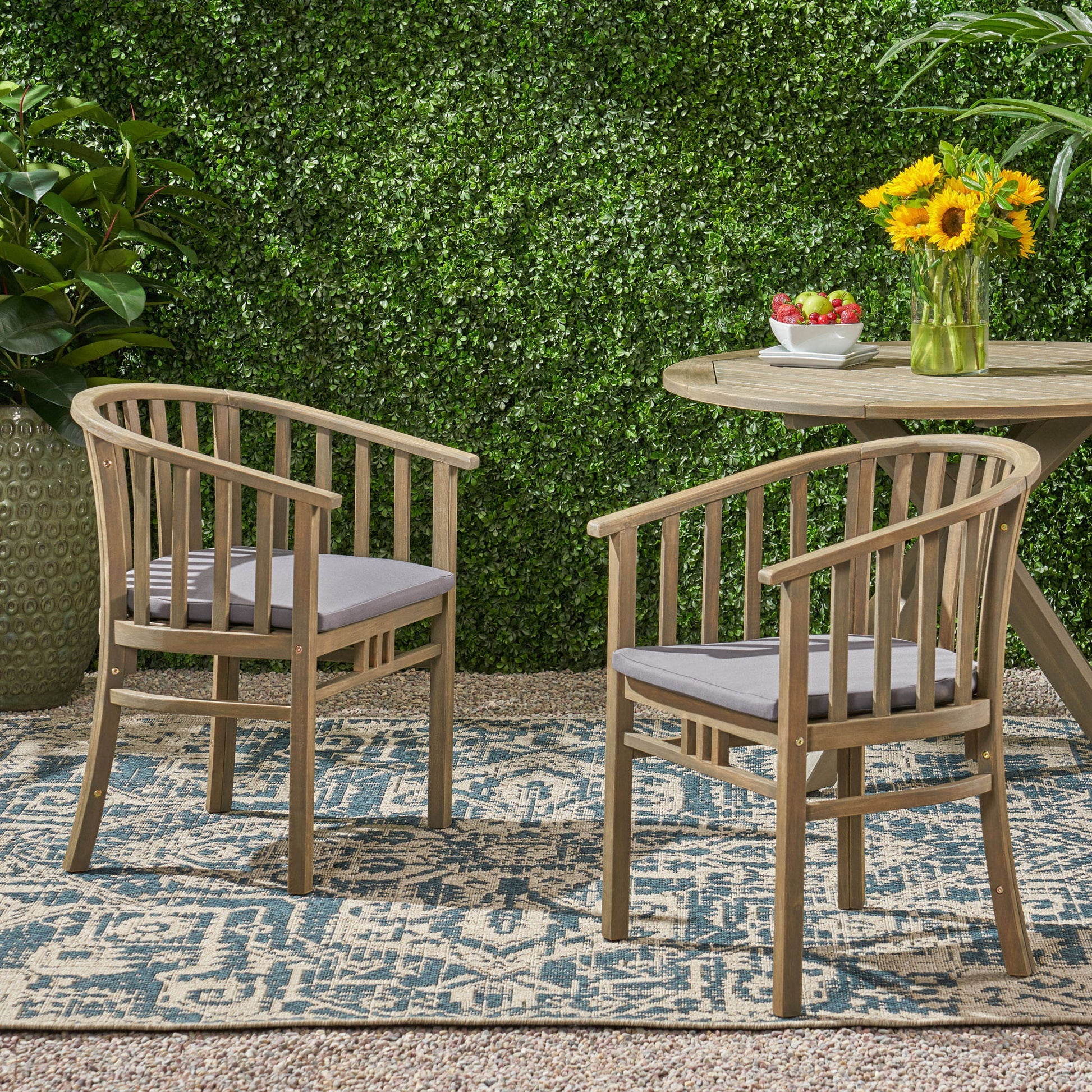 Two Outdoor Wooden Dining Chairs With Cushions (Set of 2) with gray cushions are arranged on a patterned outdoor rug near a round wooden table topped with fruit and sunflowers, surrounded by lush greenery and a leafy hedge backdrop.