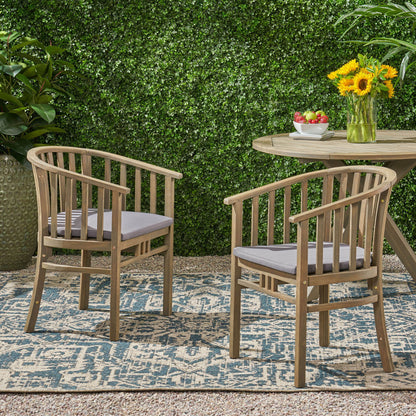 Two Outdoor Wooden Dining Chairs With Cushions (Set of 2) with gray cushions are arranged on a patterned outdoor rug near a round wooden table topped with fruit and sunflowers, surrounded by lush greenery and a leafy hedge backdrop.