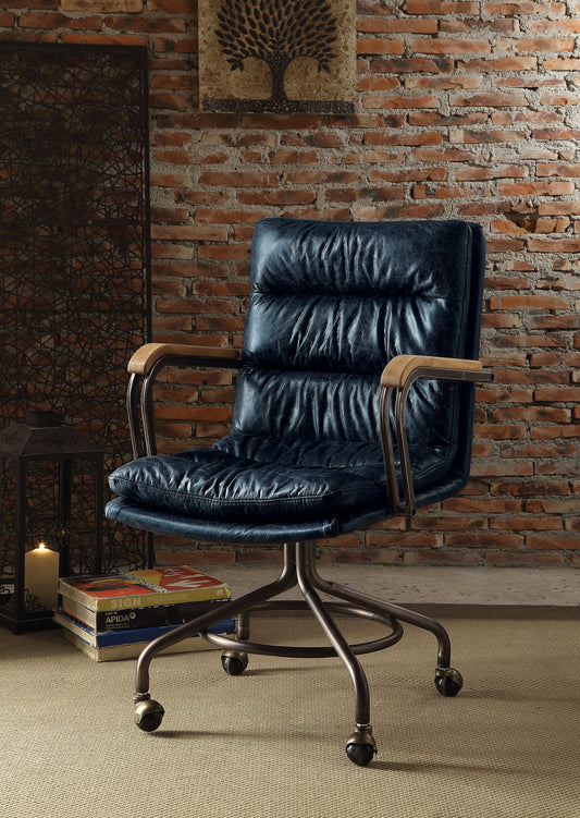 The Harith - Retro Top Grain Leather Office Chair with metal legs and wooden armrests is placed on a beige carpet before a brick wall, beside a stack of books, a candle, and decorative artwork.