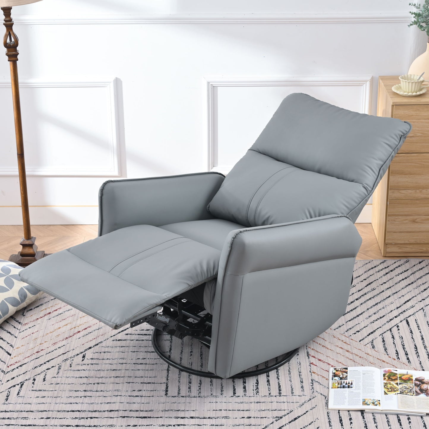The 360° Modern Small Rocking Swivel Recliner Chair With Side Pocket in gray sits on a patterned rug in a bright living room, next to a wooden nightstand, floor lamp, and an open magazine lying on the floor.