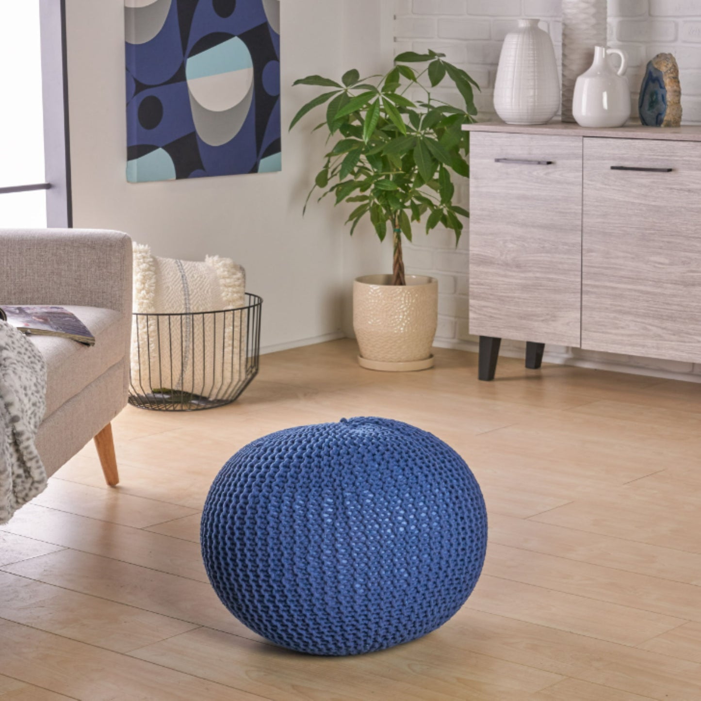 A modern living room with light wood flooring, a gray sofa, a potted plant, white vases on a cabinet, blankets in a wire basket, abstract wall art, and the Bordeaux Knitted Cotton Round Pouf used as an ottoman and floor cushion.