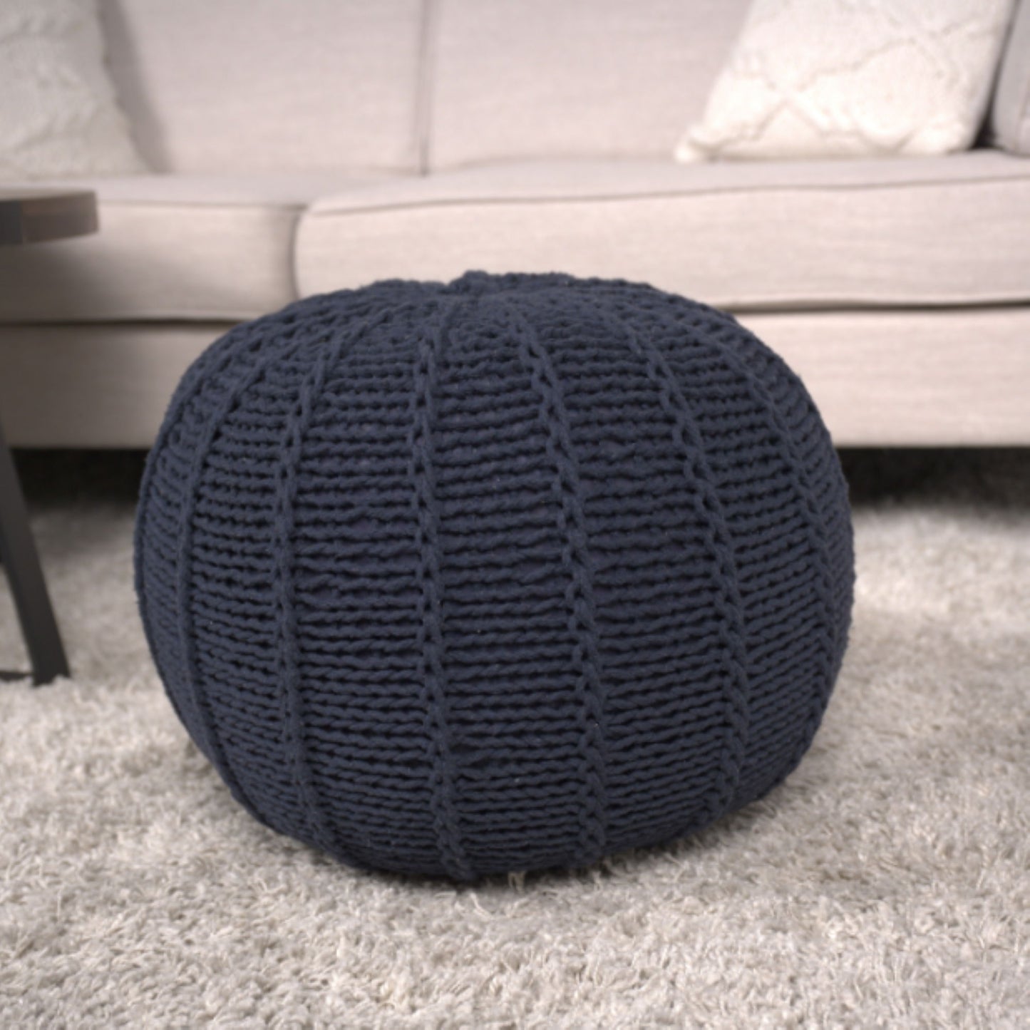 The Bordeaux Knitted Cotton Round Pouf Ottoman in dark blue rests on a light gray carpet before a beige sofa with patterned cushions, beside a dark-colored table.