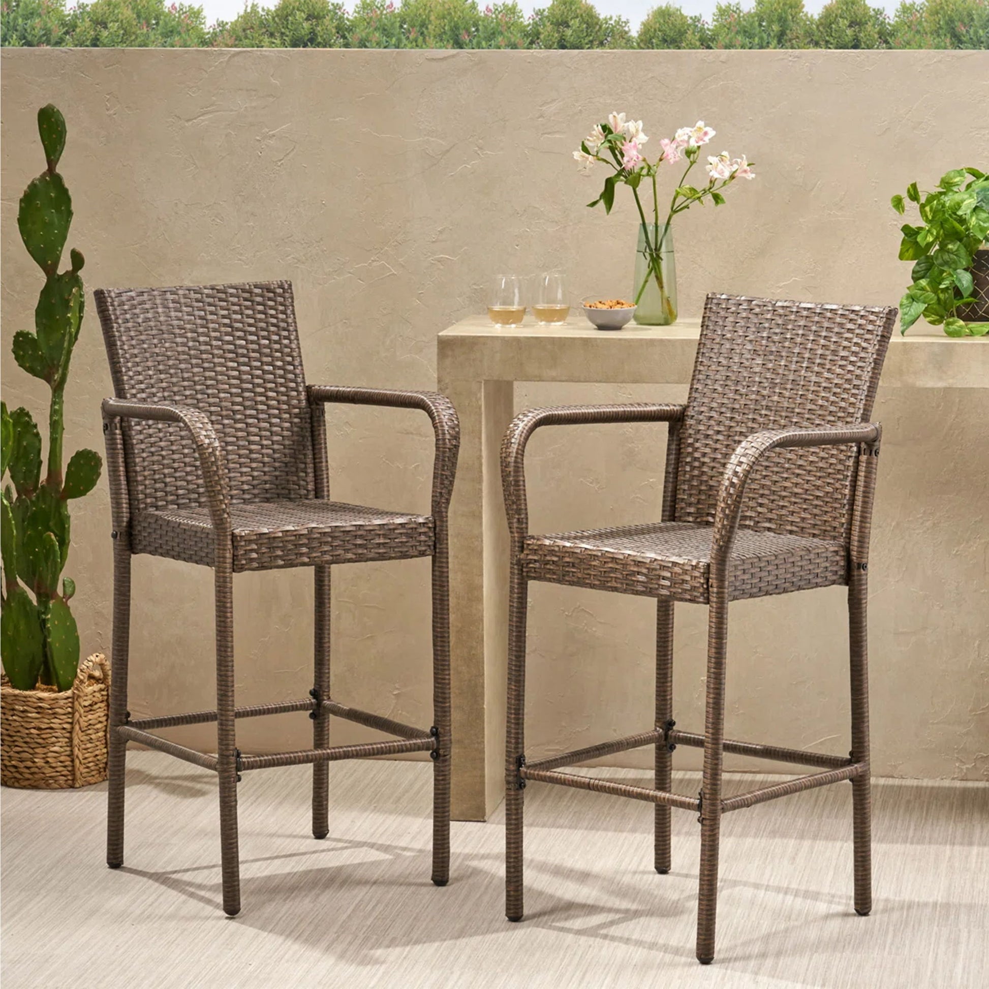 Two Outdoor Wicker Barstools with rattan and iron frames sit on a patio by a small table holding flowers, glasses, and plants; greenery and a cactus are seen in front of a beige wall with trees in the background.