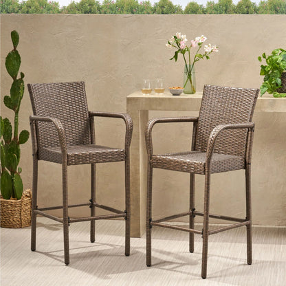 Two Outdoor Wicker Barstools with rattan and iron frames sit on a patio by a small table holding flowers, glasses, and plants; greenery and a cactus are seen in front of a beige wall with trees in the background.