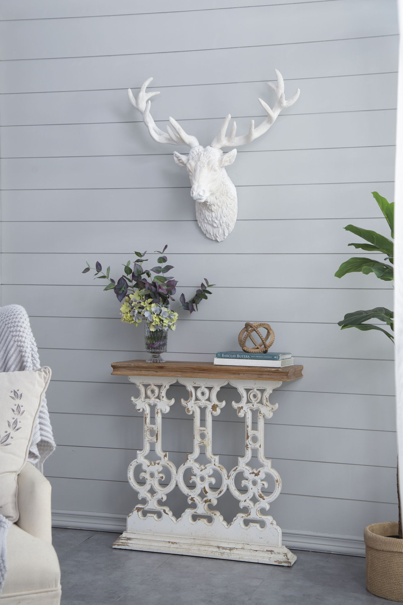 A Classic Vintage Wash Console Table With Wood Top - Brown / Antique White sits against a light gray shiplap wall, styled with books, flowers in a vase, and decor. Above, a white deer head mount hangs; a leafy plant stands nearby.