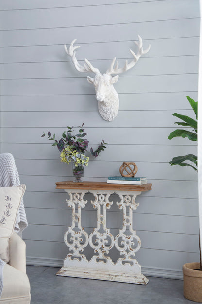 A Classic Vintage Wash Console Table With Wood Top - Brown / Antique White sits against a light gray shiplap wall, styled with books, flowers in a vase, and decor. Above, a white deer head mount hangs; a leafy plant stands nearby.