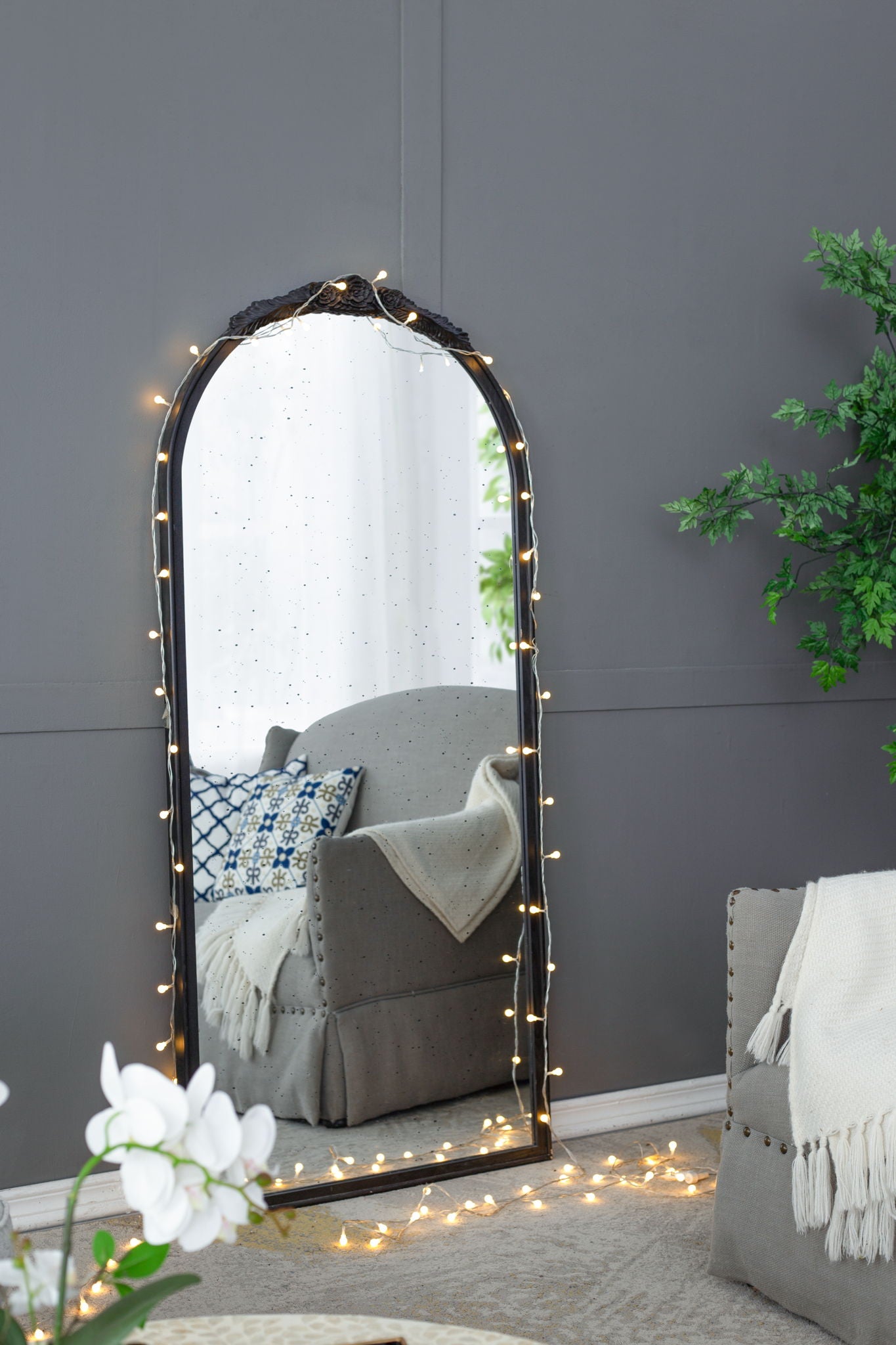 The Hand Carved Rose Antique Mirror Frame, a wood arch mirror wall decor for the living room, sits on the floor with string lights, reflecting a gray armchair with pillows and draped blanket amid a potted plant and light rug for a cozy vibe.