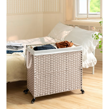 A Laundry Hamper With Lid, featuring a PE rattan frame and powder coating, with two removable bags, is placed on wheels next to a bed in a bright bedroom with light walls and natural window light.