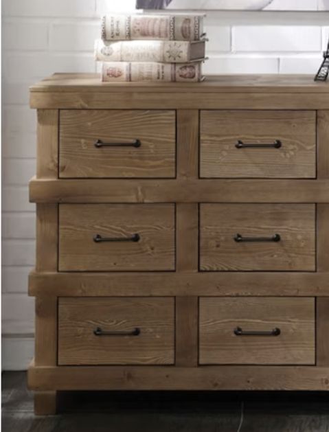 The Adams - Antique Dresser in Brown Oak features nine drawers with black metal handles. Three decoratively covered books rest on top, all set against a white brick wall background.
