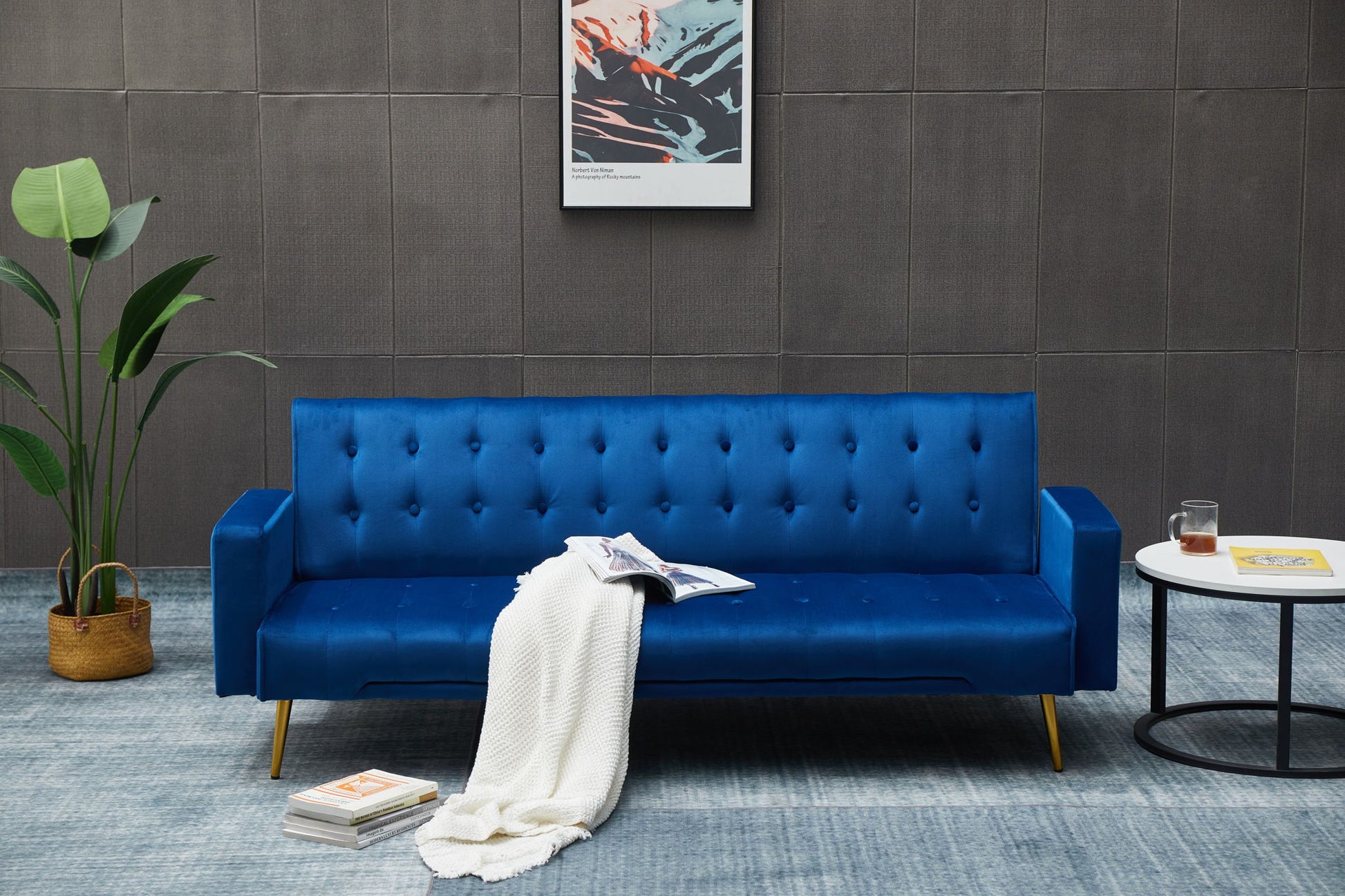 A modern living room features the Comfort Compact Design Sofa Bed in blue, topped with a white blanket, an open magazine, a potted plant nearby, a round side table with a drink, and wall art on a gray wall.