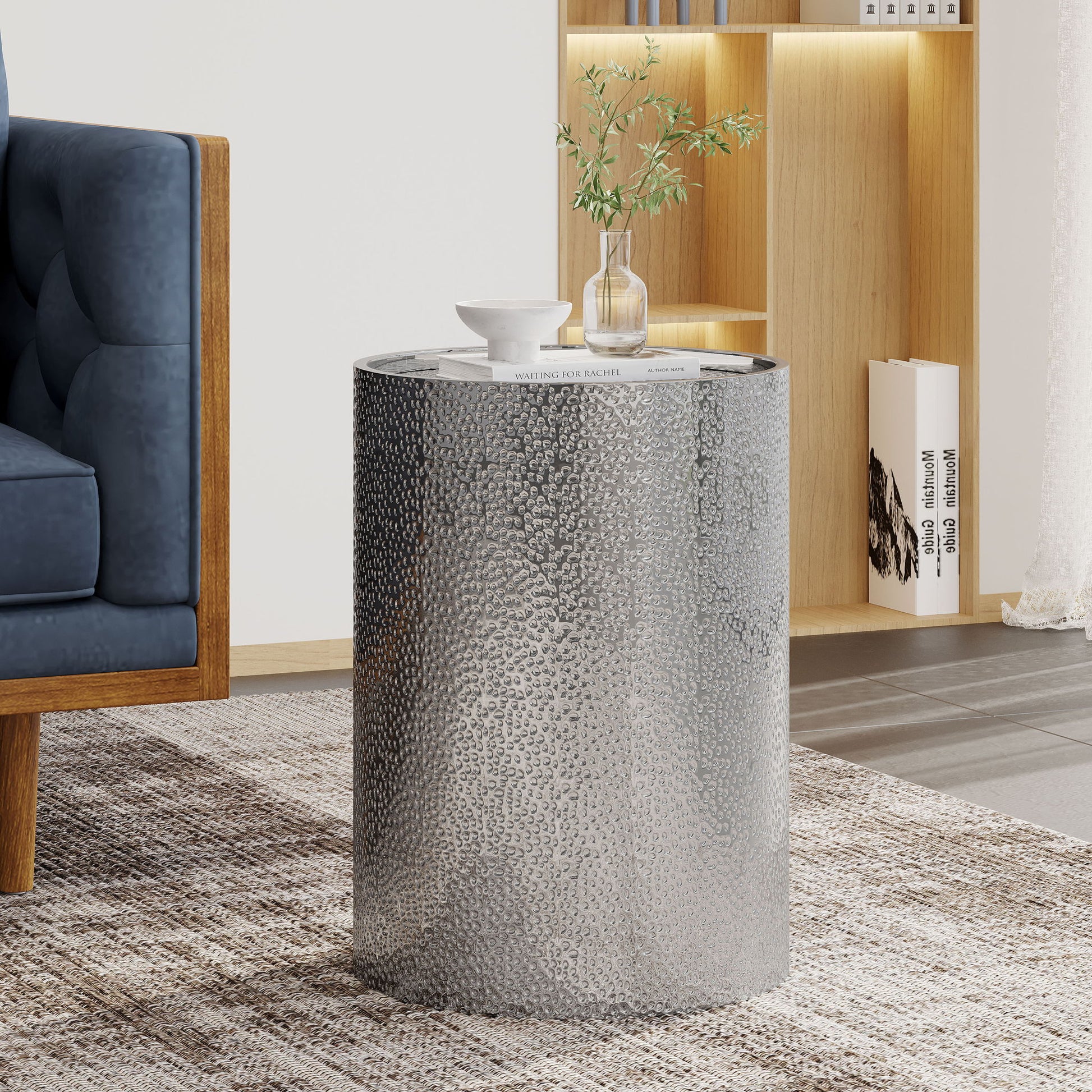 A Modern Round Accent Table With Hammered Surface sits on a patterned rug beside a blue sofa, topped with a small vase of greenery, a white cup, and a book. A wooden bookshelf is visible in the background.