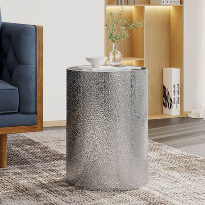 A Modern Round Accent Table With Hammered Surface sits on a patterned rug beside a blue sofa, topped with a small vase of greenery, a white cup, and a book. A wooden bookshelf is visible in the background.
