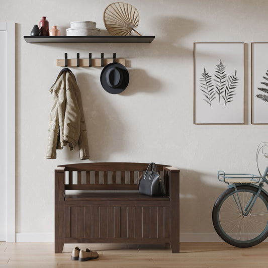 A cozy entryway features the Acadian - Entryway Storage Bench, Handcrafted, with shoes and a handbag atop. Above, a coat rack holds a jacket and hat. A shelf displays decor, framed botanical prints hang nearby, and part of a bicycle is visible.