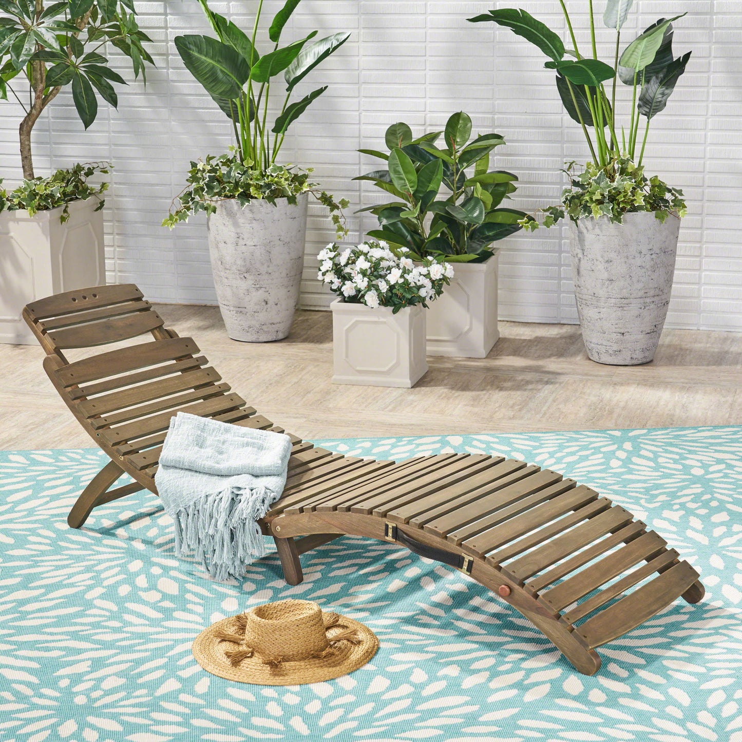 The Lahaina - Wood Foldable Chaise Lounge, topped with a folded blue blanket and straw hat, rests on a blue patterned rug amid large potted green plants against a white brick wall.