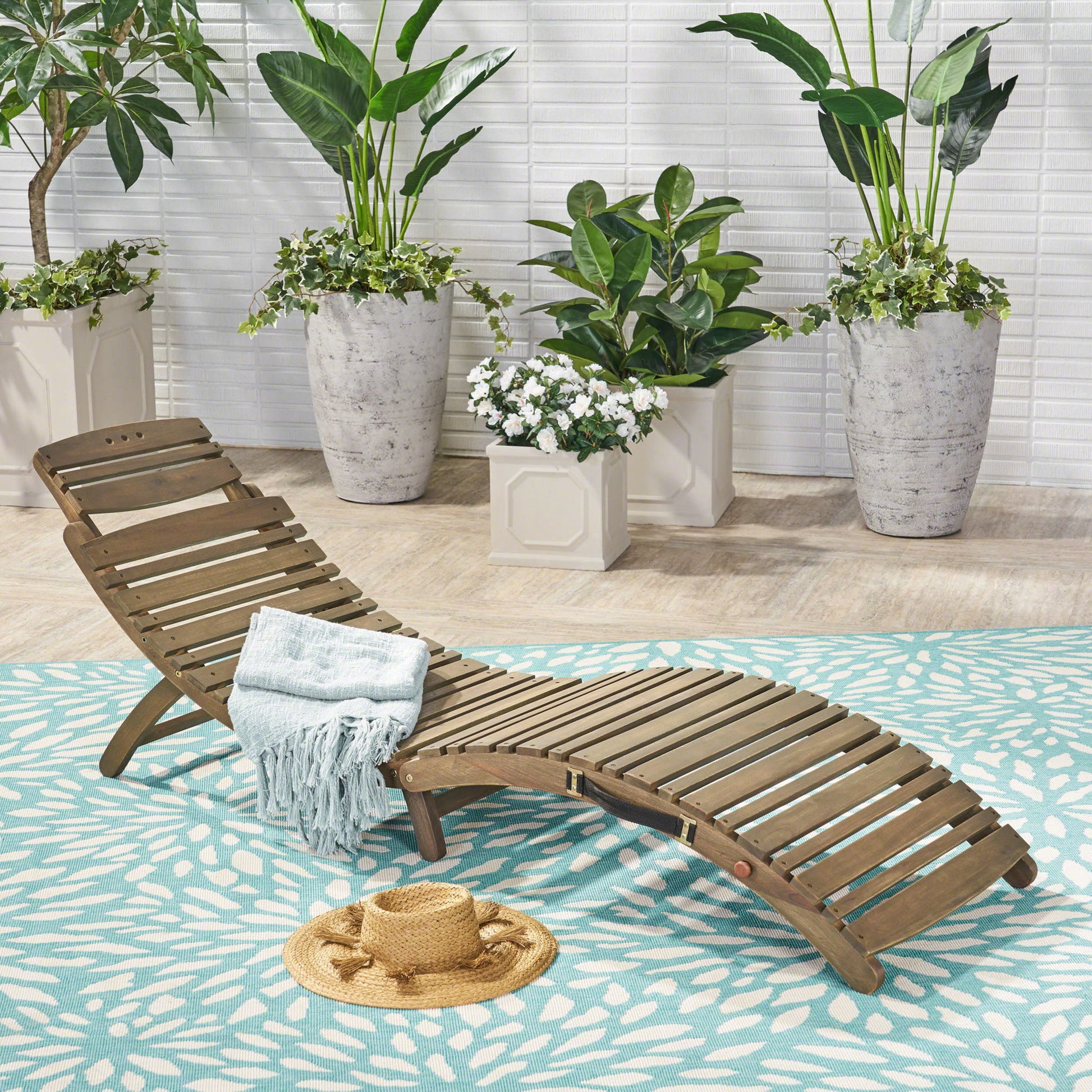The Lahaina - Wood Foldable Chaise Lounge, topped with a folded blue blanket and straw hat, rests on a blue patterned rug amid large potted green plants against a white brick wall.