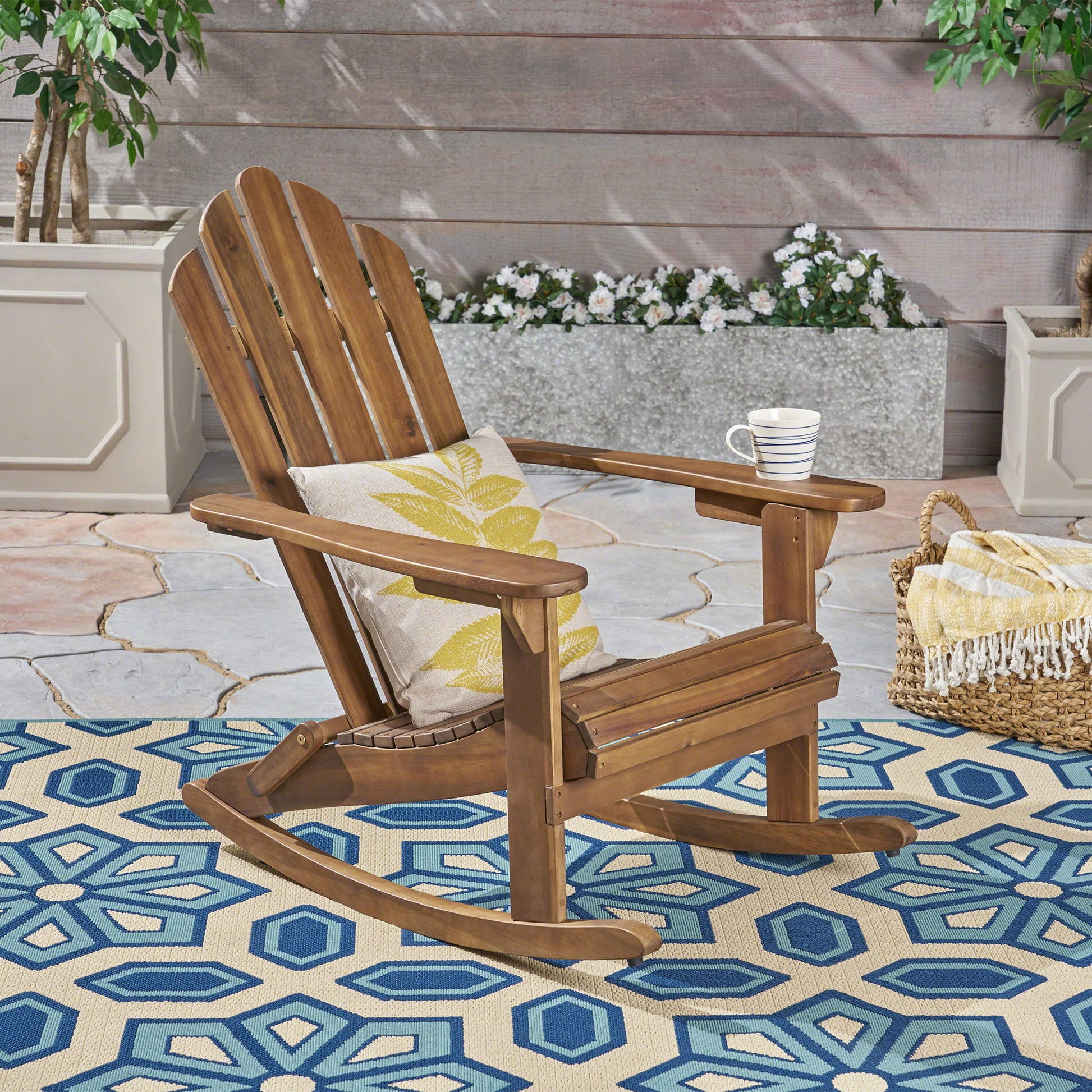 The Echoes Outdoor Acacia Wood Adirondack Rocking Chair sits with a pillow and mug on its armrest atop a blue geometric outdoor rug, surrounded by potted plants and flowers on a stone patio.