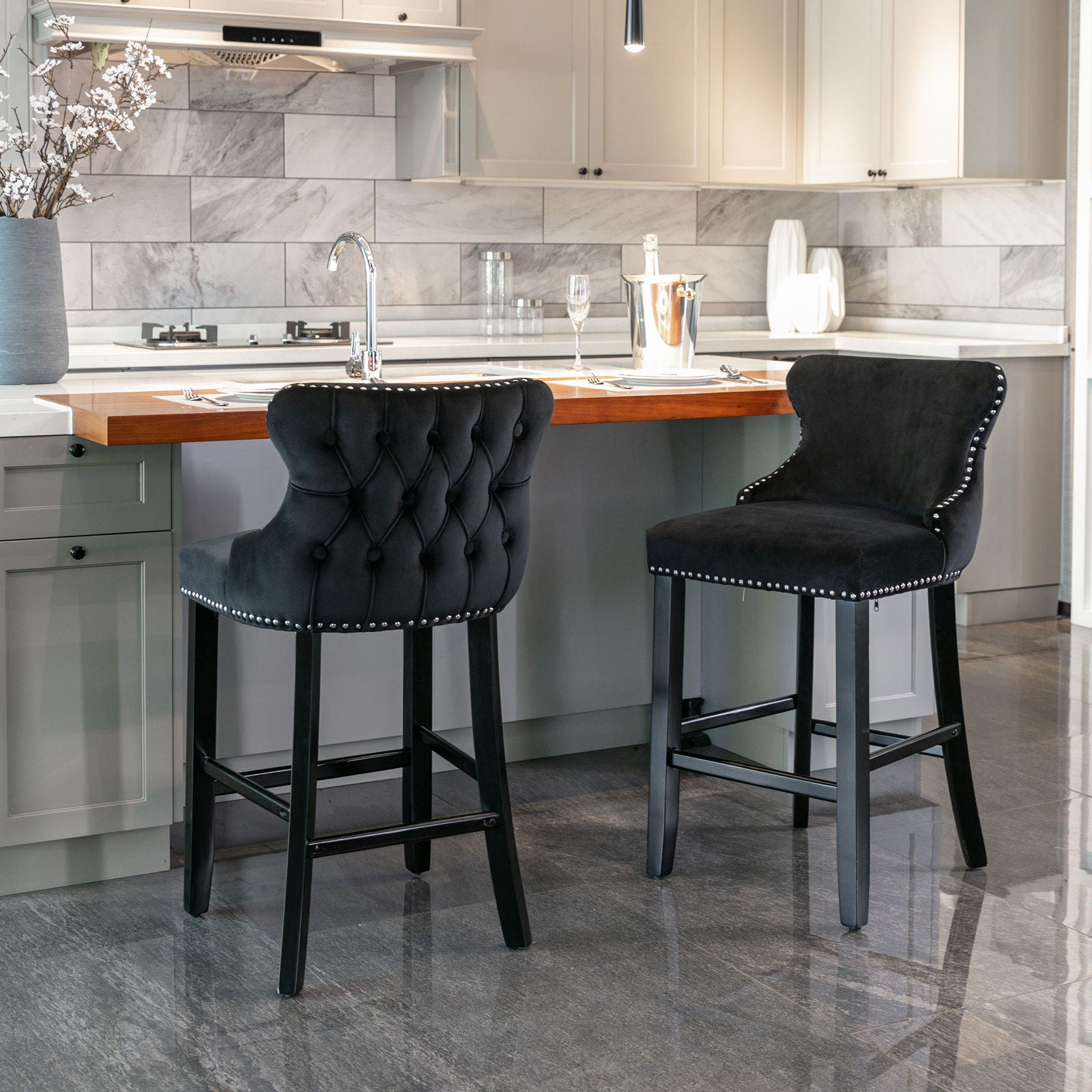 A set of two Contemporary Velvet Upholstered Wing-Back Barstools with button tufted backs, wooden legs, and chrome nailhead trim is placed at a kitchen island with a wood countertop in a modern kitchen with gray cabinets and a marble backsplash.