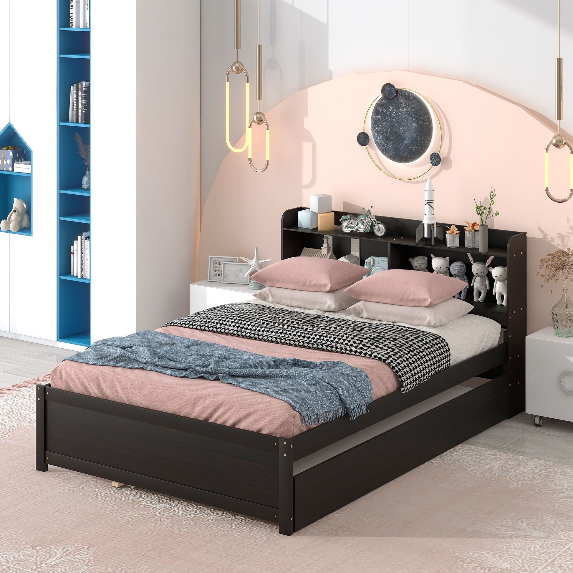 A modern bedroom features the Full Bed With Trundle, Bookcase in dark wood, pink and gray bedding with pillows, a blue textured throw, and a built-in headboard shelf holding books and stuffed animals. Pendant lights and wall art hang above.