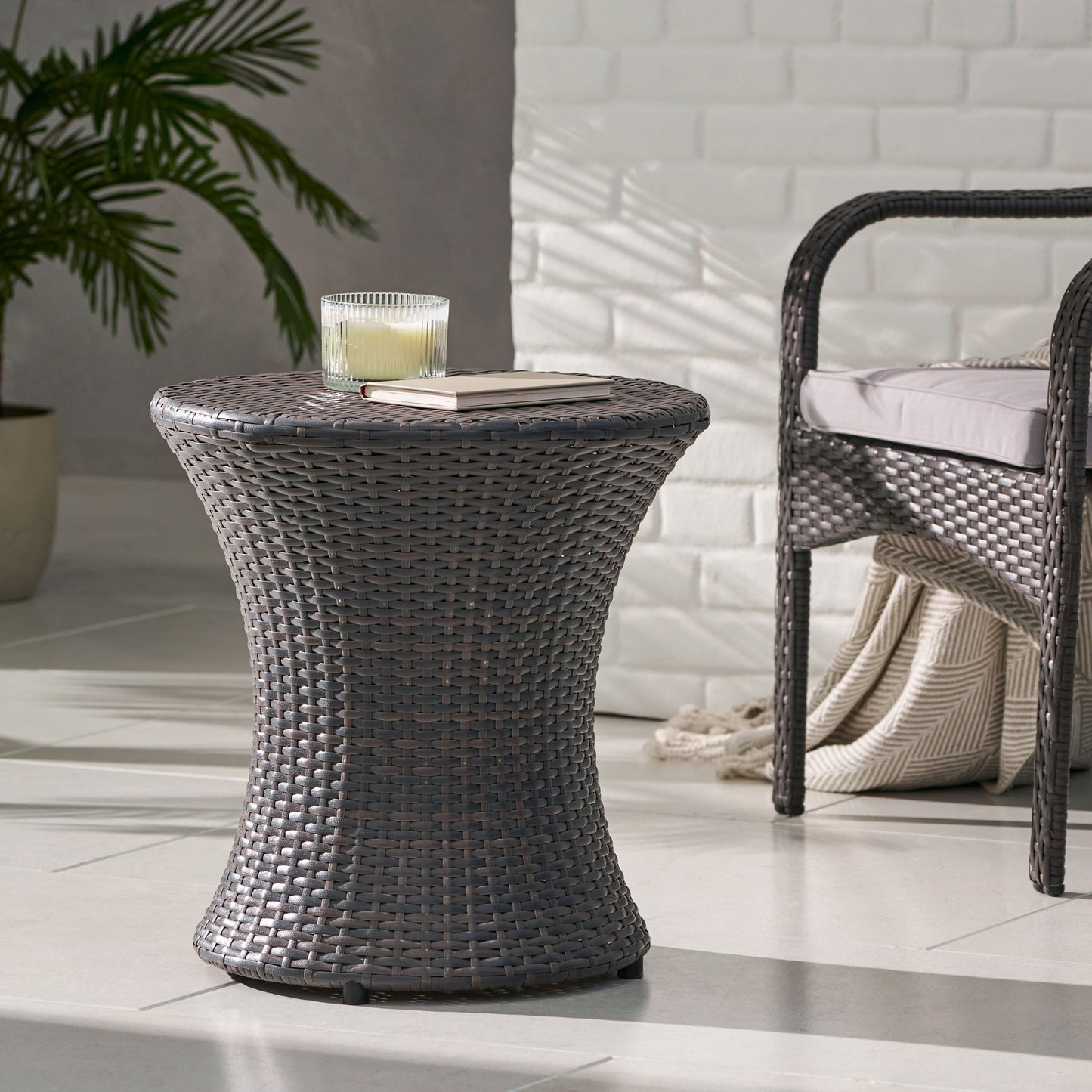 A Thrive Hourglass Rattan Table (no glass top) with a candle and closed book sits beside a wicker chair with beige cushion and folded blanket, on tiled flooring with a potted plant in the background.