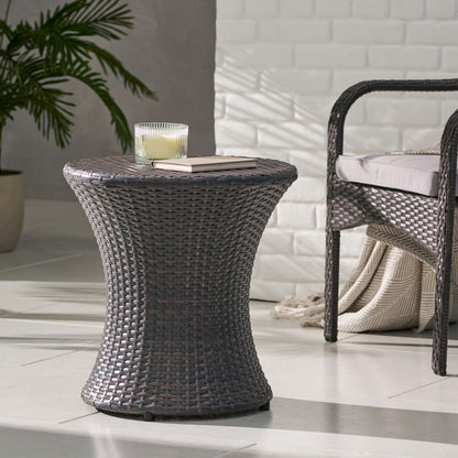 A Thrive Hourglass Rattan Table (no glass top) with a candle and closed book sits beside a wicker chair with beige cushion and folded blanket, on tiled flooring with a potted plant in the background.