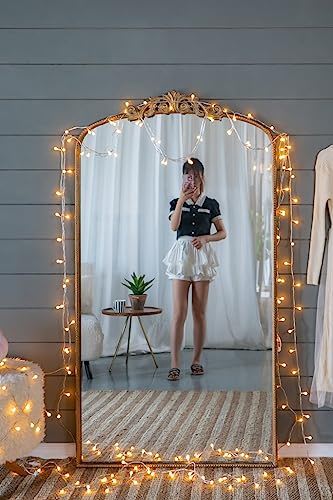 A woman takes a mirror selfie in front of the Arch Mirror, Baroque Inspired Wall Decor, with string lights draped over it. She wears a black top and white skirt; behind her are sheer curtains, a small table with a potted plant, and a hanging dress.