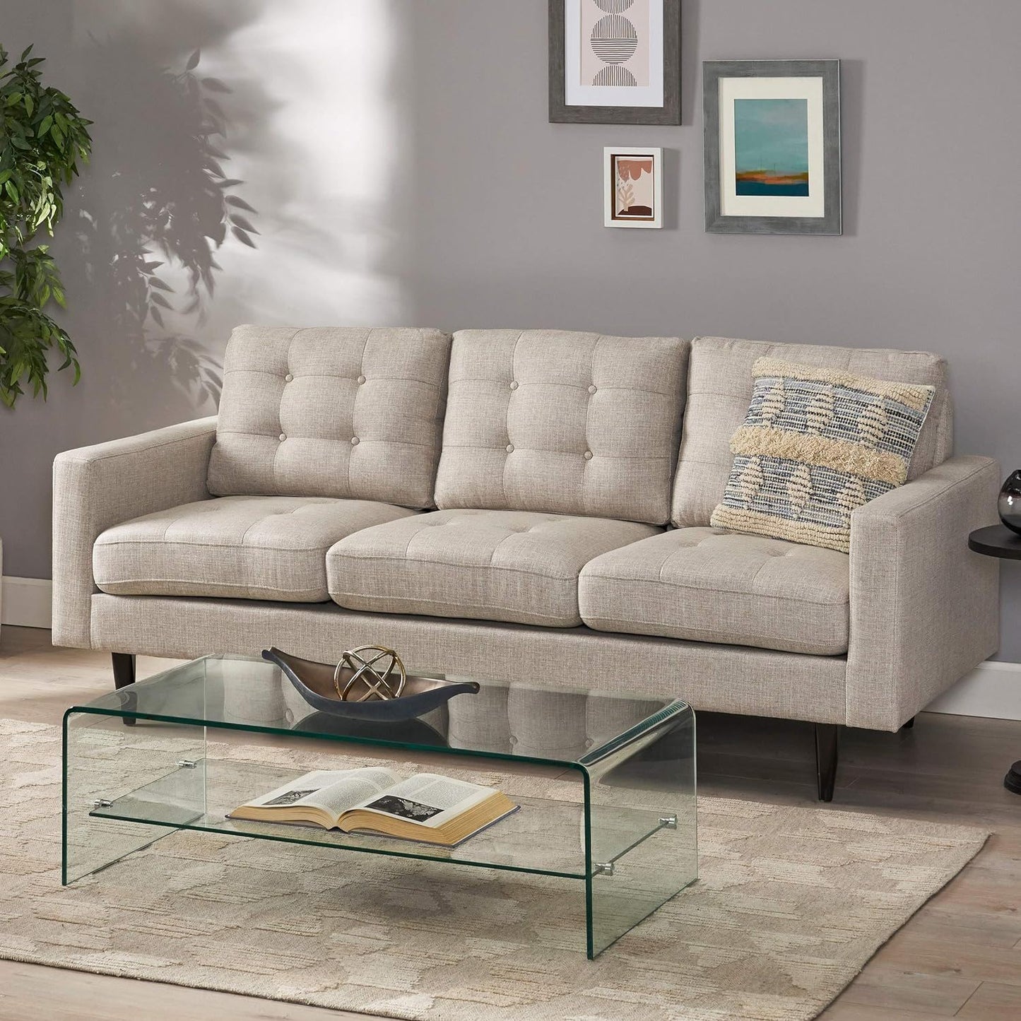 A modern living room featuring the Jenny Contemporary Tufted Fabric 3 Seater Sofa in light beige, a patterned pillow, glass coffee table with book and tray, beige rug, framed art on gray walls, and a potted plant in the corner.