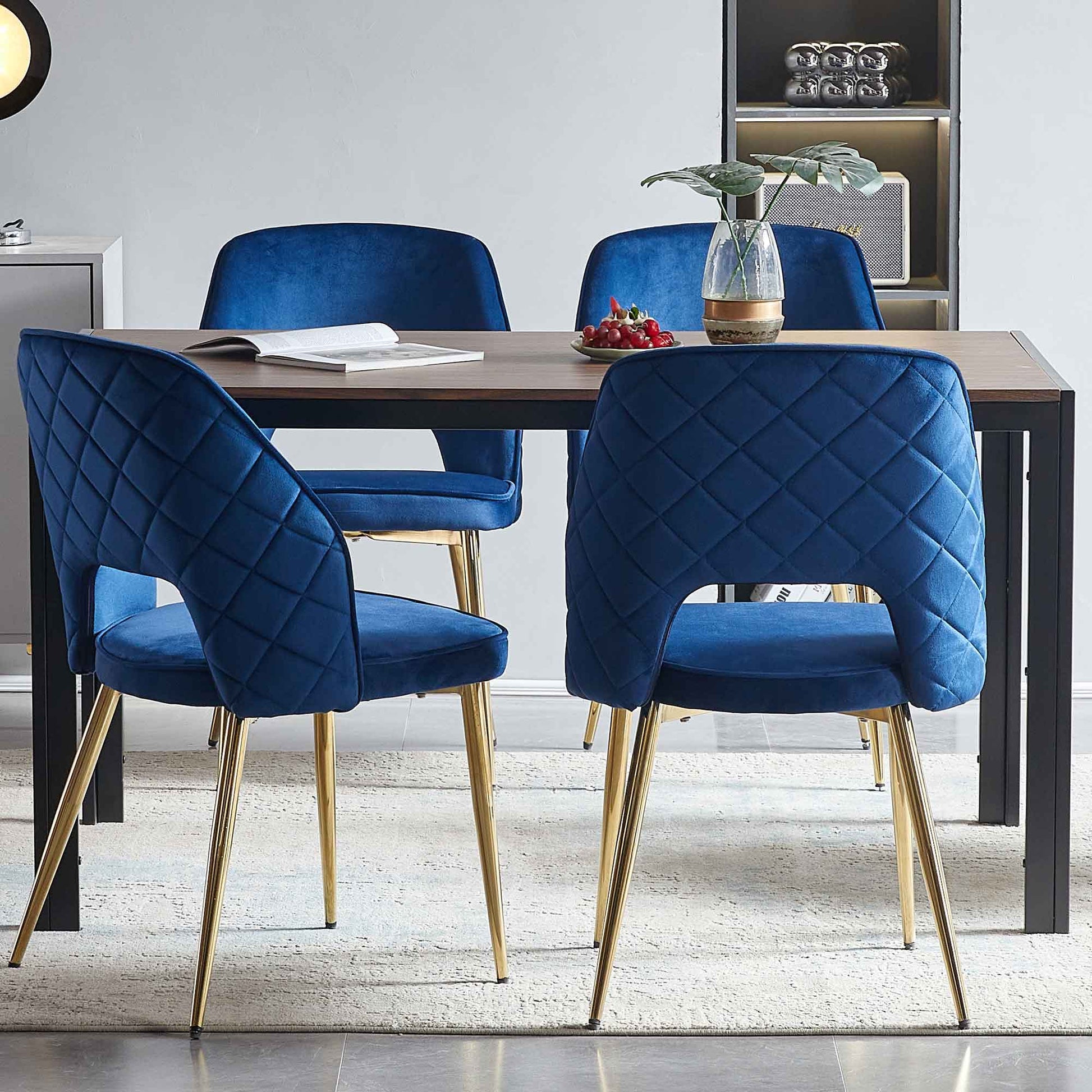 A modern dining area features a rectangular wooden table, four velvet dining chairs with metal legs and hollow backs, a beige rug, an open book, a vase with leaves, and a plate of cherries.