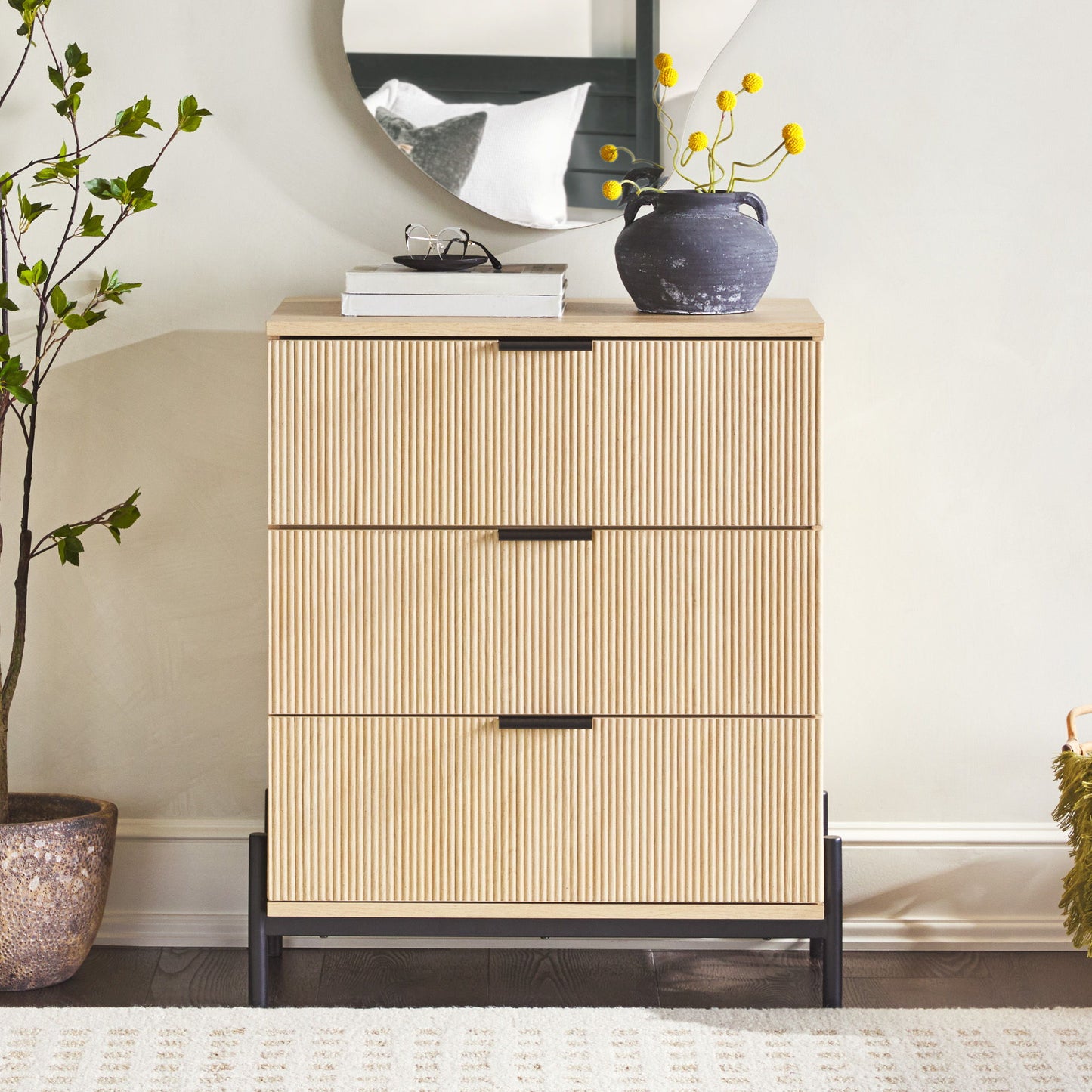 The Mid-Century 3 Drawer Chest With Reeded Drawer Fronts stands against a light wall, topped with a round mirror, potted plant, and vase of yellow flowers. A leafy plant is positioned to the left.