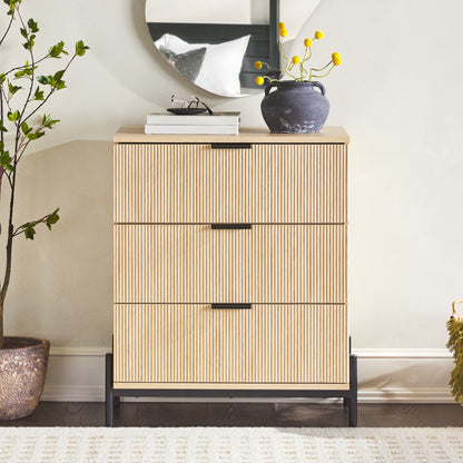 The Mid-Century 3 Drawer Chest With Reeded Drawer Fronts stands against a light wall, topped with a round mirror, potted plant, and vase of yellow flowers. A leafy plant is positioned to the left.