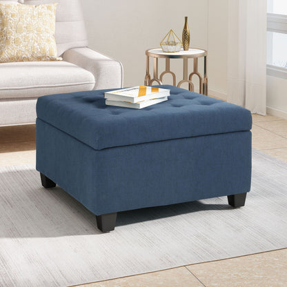 The Isabella - Contemporary Ottoman With Storage, in blue upholstery with a tufted top, sits on a light area rug in a bright living room. Two books are stacked on it, near a beige sofa, patterned pillow, and round side table with décor.