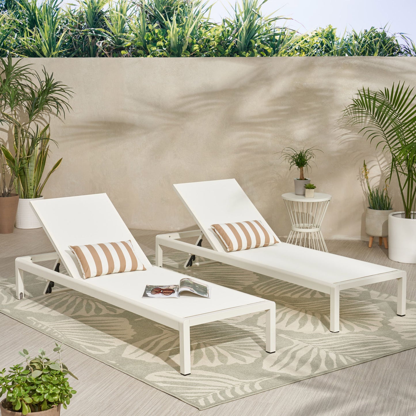 Two Outdoor Patio Chaise Lounges with 5 Reclining Positions, each with a striped pillow, sit on a leaf-patterned rug in a bright outdoor area with potted plants. An open magazine and sunglasses rest on one chaise near a light wall.