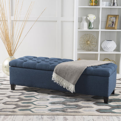 The Glouster Contemporary Storage Ottoman with button tufted design sits on a geometric rug in a modern living room, topped with a gray fringed throw blanket and backed by a white shelving unit with decorative items.