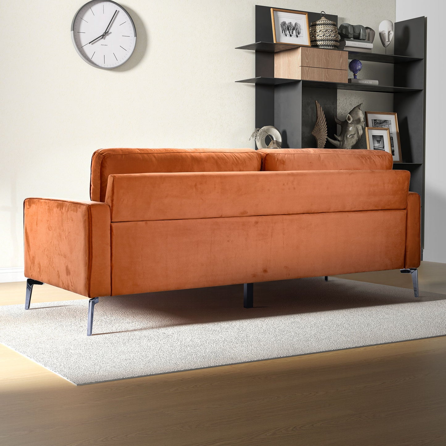 The Axten - Sofa - Orange with metal legs sits on a light rug in a living room with a wood floor, backed by a wall clock and black shelves holding books, photos, and decor.