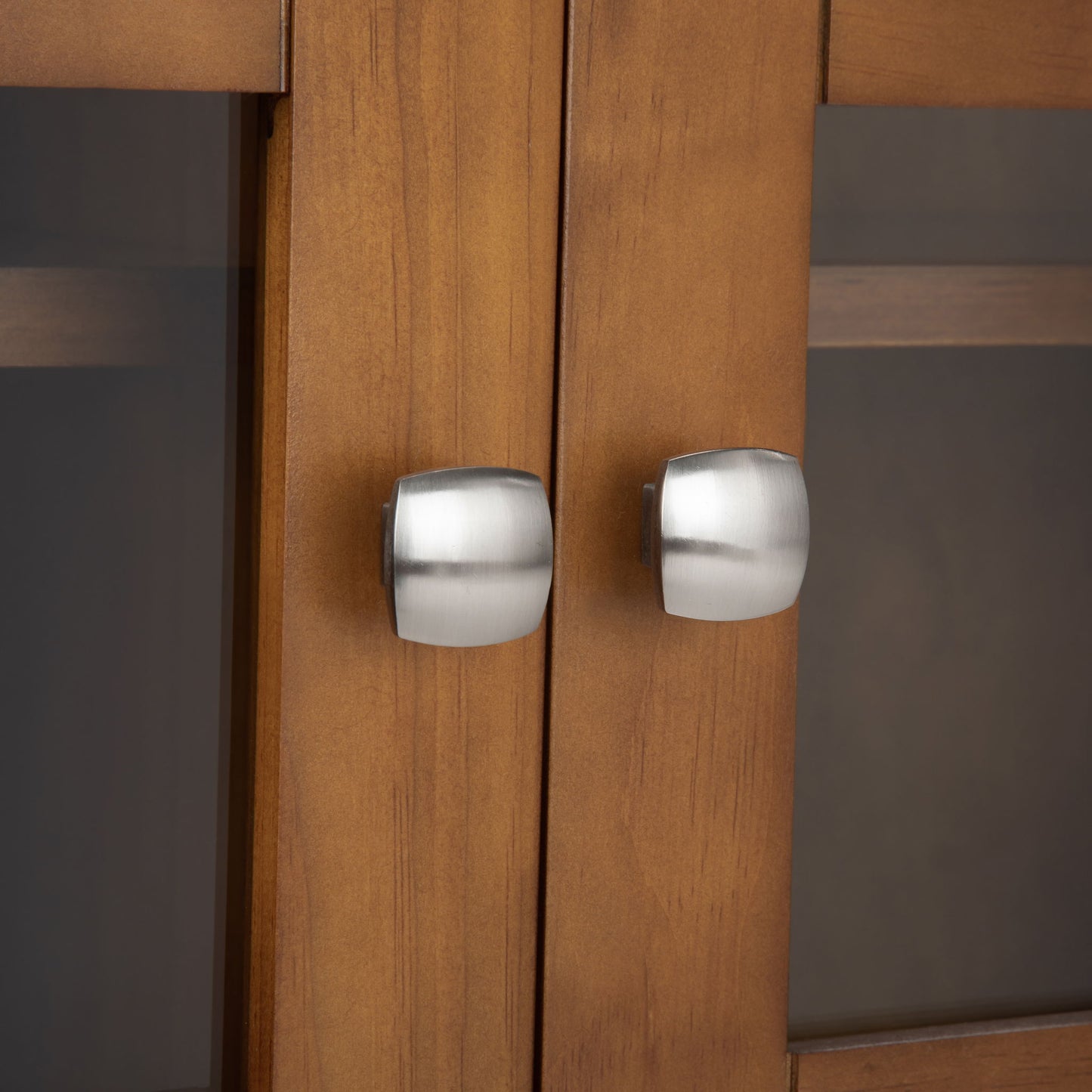 Close-up of two brushed metal, modern square knobs on the Acadian - Medium Storage Cabinet, Handcrafted. The cabinet features light brown wood and glass panel doors for a sleek, contemporary look.