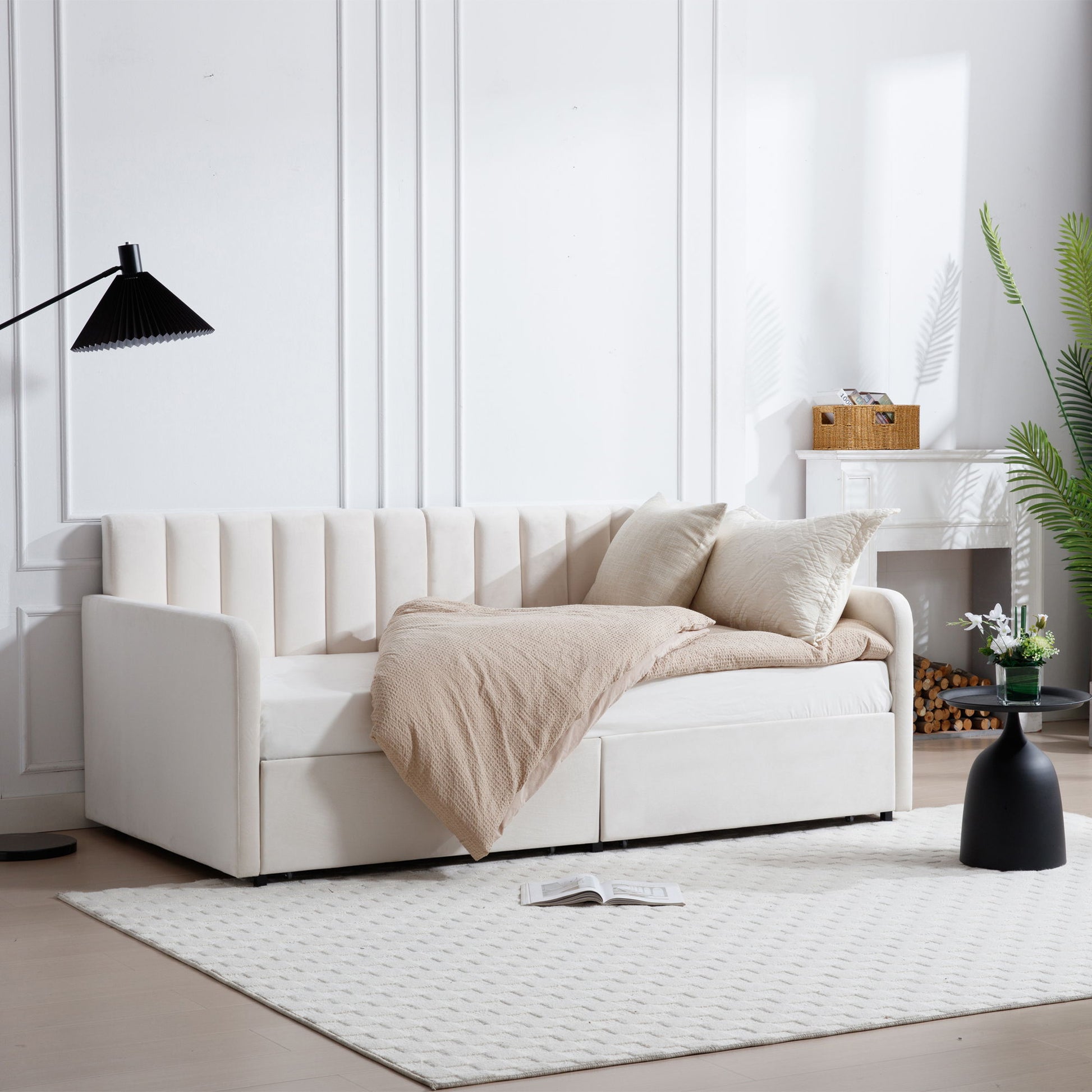 The Flora Upholstered Daybed with a ribbed tufted backrest sits in a bright living room, styled with a beige blanket and pillows. A black floor lamp, potted plants, and an open book add to the cozy minimalist ambiance.