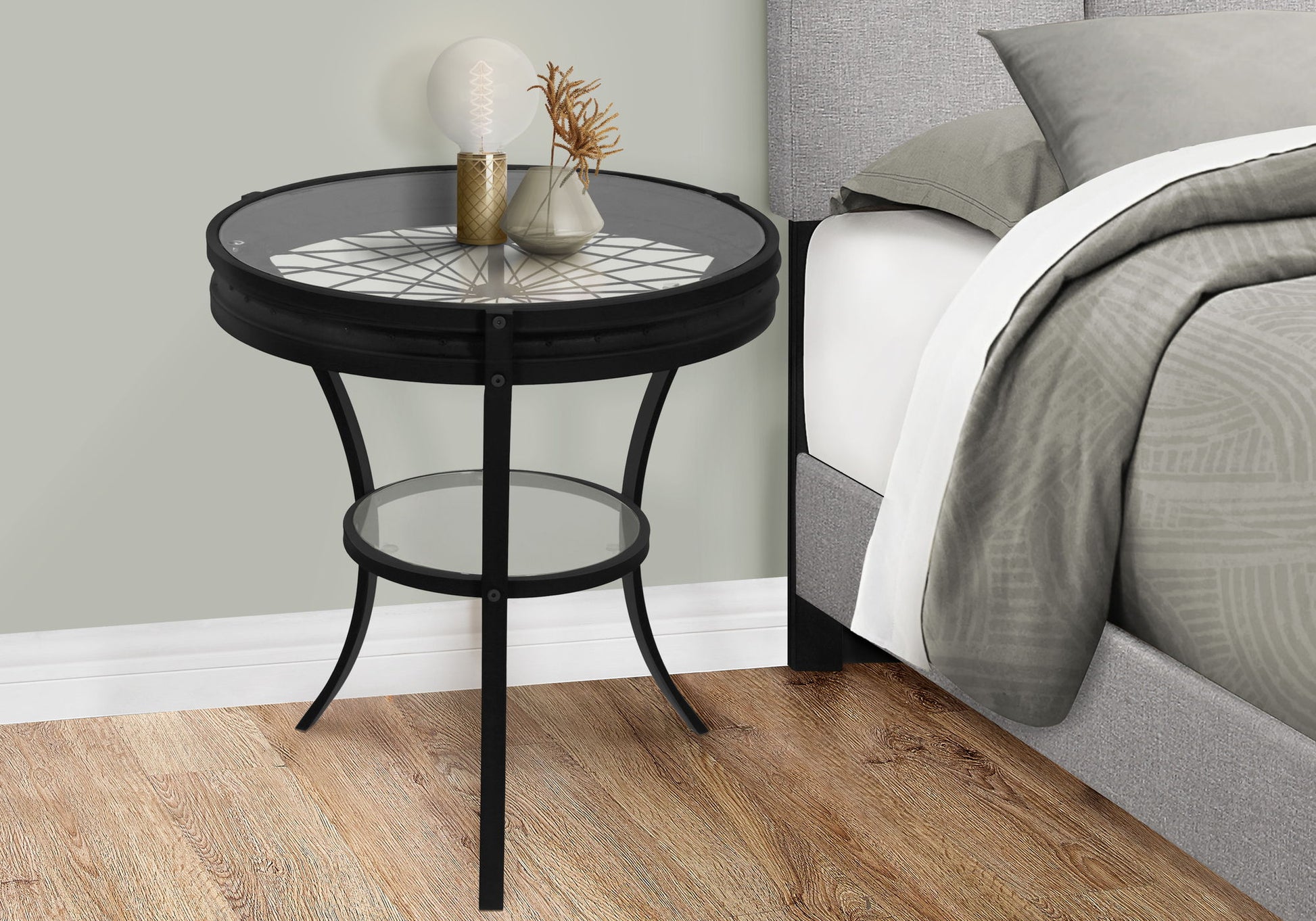 A Round Accent Table, Transitional - Black with a patterned glass top and lower shelf stands by a gray upholstered bed dressed in beige and gray bedding. The table displays a decorative lamp, gold vase, and glass vase with dried foliage.