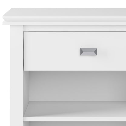 A close-up of the Artisan - Handcrafted Bedside Table shows a white, modern nightstand with one drawer featuring a rectangular silver handle and an open shelf below. Its minimalistic design adds elegance to any space.