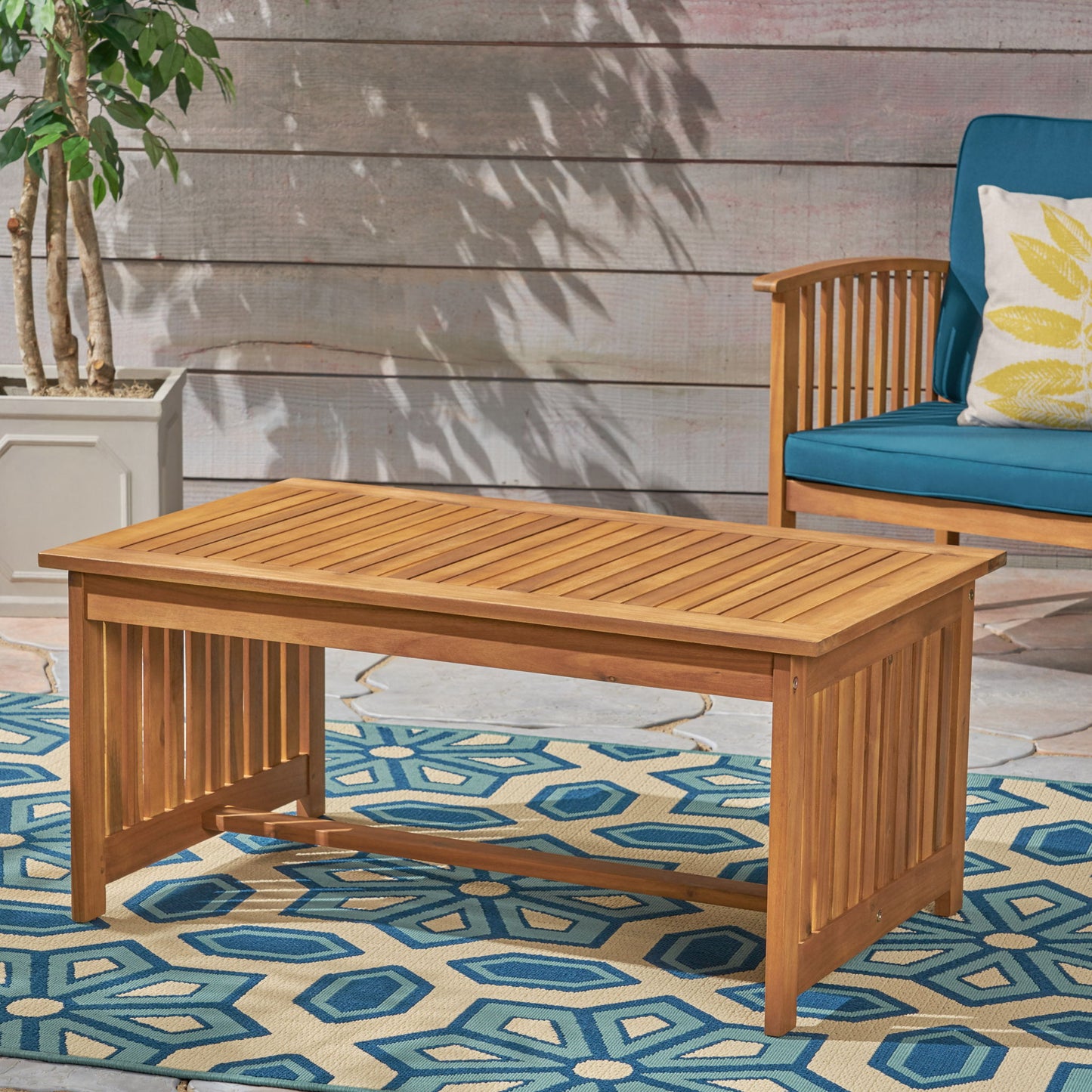An Acacia Wood Rectangular Patio Coffee Table for outside sits on a blue geometric patterned rug, beside a potted plant and a wooden chair with blue cushions and a yellow patterned pillow.