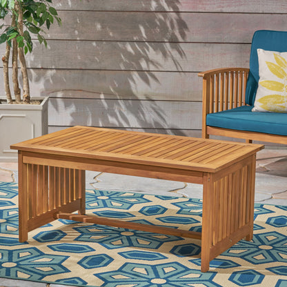An Acacia Wood Rectangular Patio Coffee Table for outside sits on a blue geometric patterned rug, beside a potted plant and a wooden chair with blue cushions and a yellow patterned pillow.