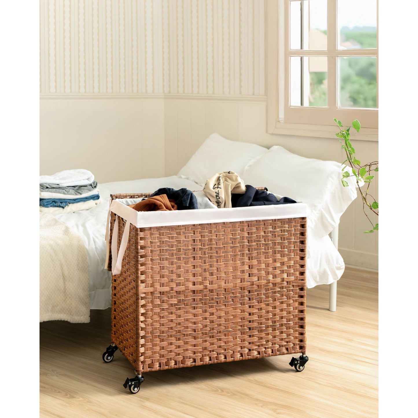 The Laundry Hamper With Lid features a PE rattan design, powder coating frame, and two removable bags. It stands on caster wheels next to a bed in a bright bedroom with wooden floors and a window.