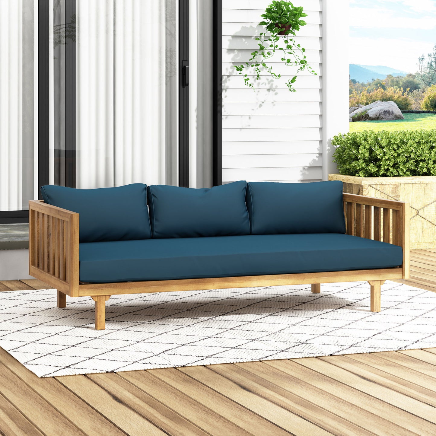 The Claremont 3 Seater Daybed, crafted from teak acacia wood with water-resistant blue cushions, is placed on a white patterned rug atop a wooden deck, set against glass doors and lush outdoor greenery.